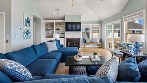 A modern living room with a sectional sofa, a fireplace, a television, and a dining area with a table and chairs. - 5514- Sisterly Love - Luxury Oceanfront | Pool, Hot Tub & Views (Nags Head)