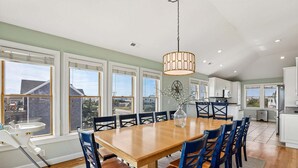 A spacious dining area with a large wooden table and blue chairs, a hanging light fixture, and large windows offering a view of the outside. - 5514- Sisterly Love - Luxury Oceanfront | Pool, Hot Tub & Views (Nags Head)