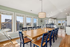 A spacious dining area with a large wooden table and blue chairs, a hanging light fixture, and large windows offering a view of the outside.