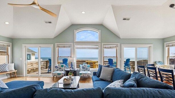 A spacious living room with a large blue sofa, a dining area with chairs, and a view of the beach through large windows. - 5514- Sisterly Love - Luxury Oceanfront | Pool, Hot Tub & Views (Nags Head)