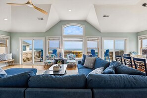 A spacious living room with a large blue sofa, a dining area with chairs, and a view of the beach through large windows. - 5514- Sisterly Love - Luxury Oceanfront | Pool, Hot Tub & Views (Nags Head)