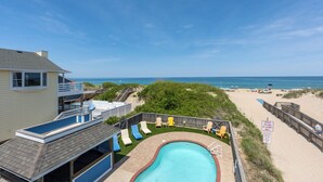 A pool area with a view of the beach and ocean, surrounded by a wooden deck and lounge chairs. - 5514- Sisterly Love - Luxury Oceanfront | Pool, Hot Tub & Views (Nags Head)