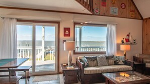 Un salon chaleureux avec un canapé, une table basse et une cheminée. Il y a deux fenêtres donnant sur la mer et un balcon avec une balustrade.