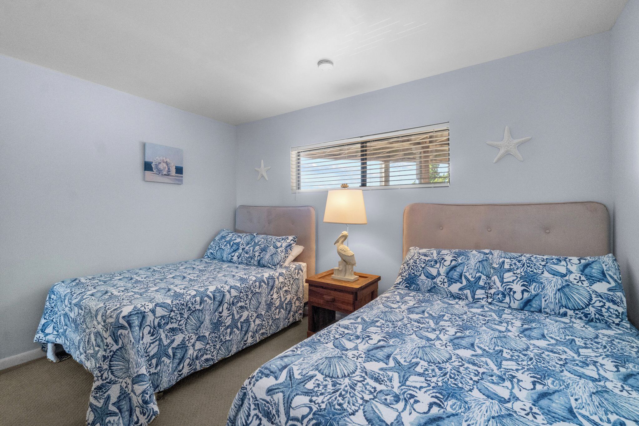 A bedroom with two beds, each with a blue floral bedspread, a nightstand with a lamp, and a window with blinds.