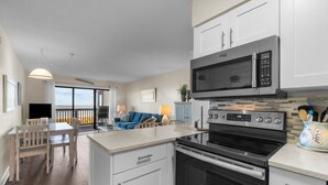 A modern kitchen with white cabinets, a black microwave, and a stainless steel oven. There is a dining area with a wooden table and chairs. - 3196 - Sea Sights - Oceanfront Duck Condo | Walk to Duck Village (Duck)
