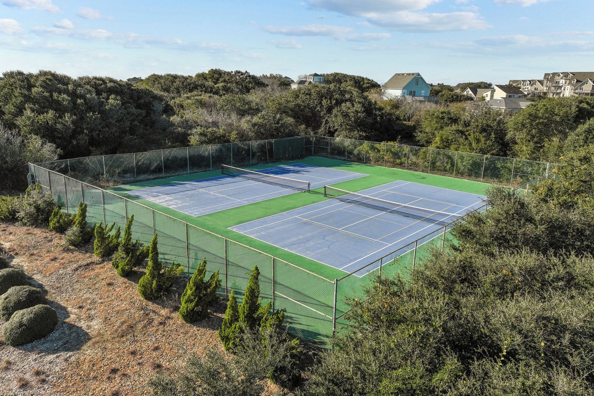 A tennis court with two courts, surrounded by a fence, and a backdrop of trees and houses.