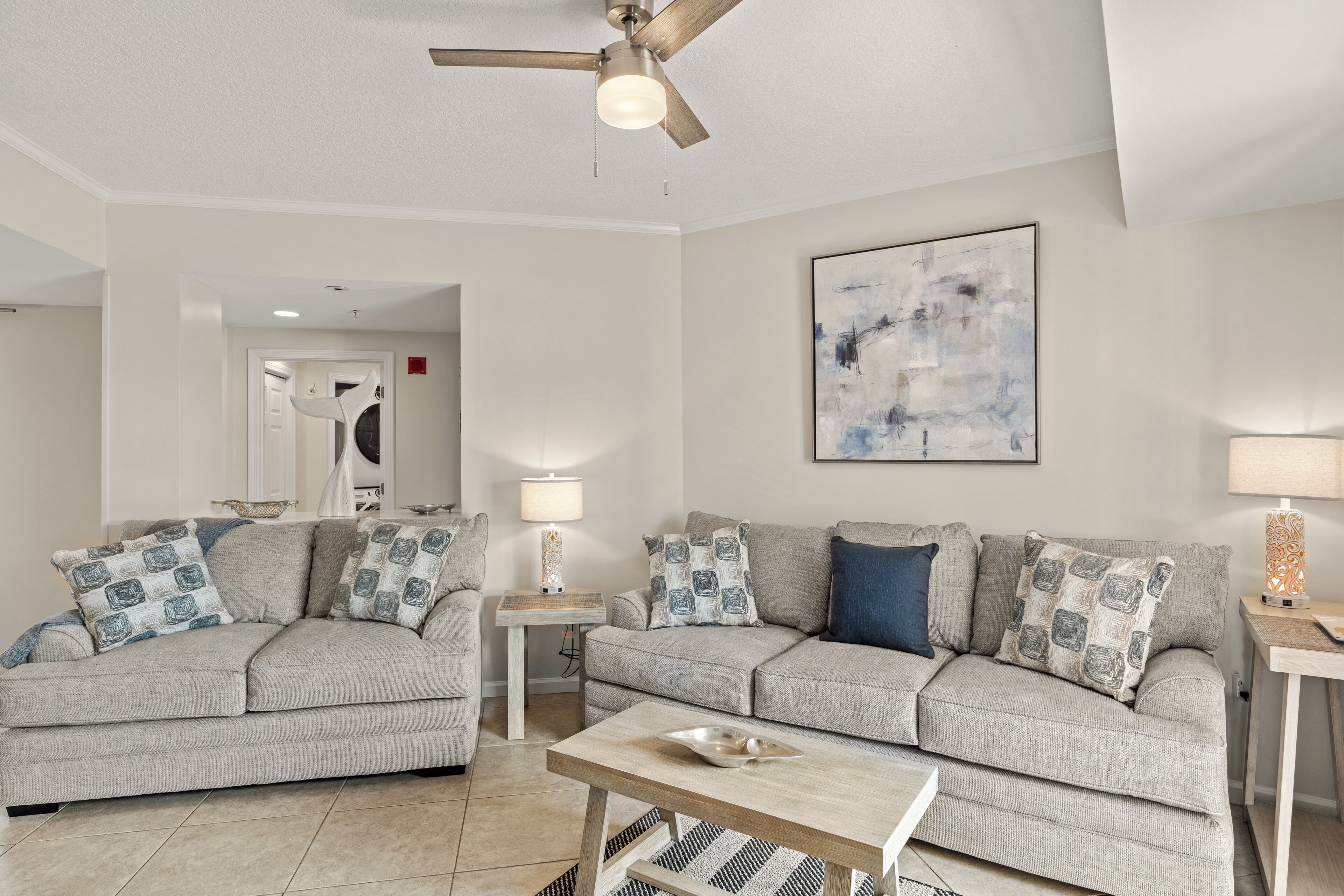 A living room with two grey sofas, a wooden coffee table, a ceiling fan, a wall-mounted abstract painting, and a side table with a lamp.