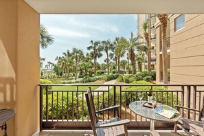 A balcony with a table, chairs, and a view of a landscaped garden with palm trees.