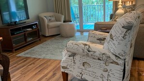 Una sala de estar con televisor, sofá, sillón y balcón con cortinas.