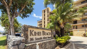 A sign for 'Kihei Beach' at 36 South Kihei Road, indicating a beachfront location with a multi-story building in the background.