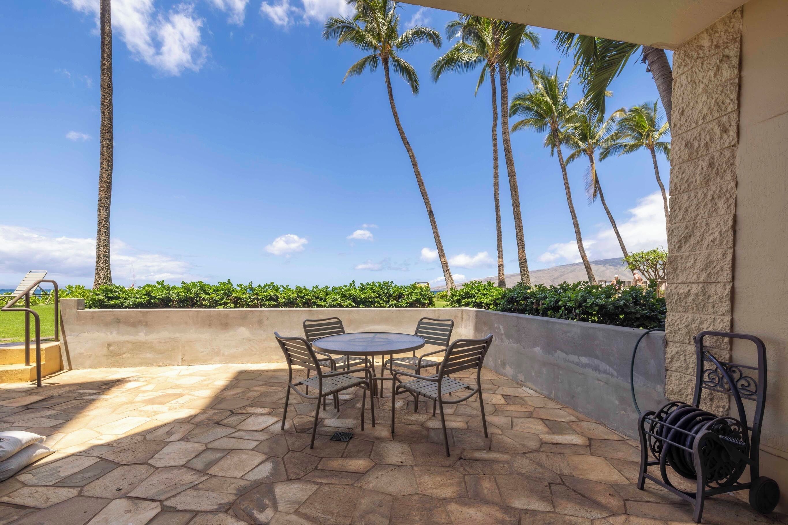 An outdoor patio with a stone floor, a round table with chairs, and a view of palm trees and mountains.