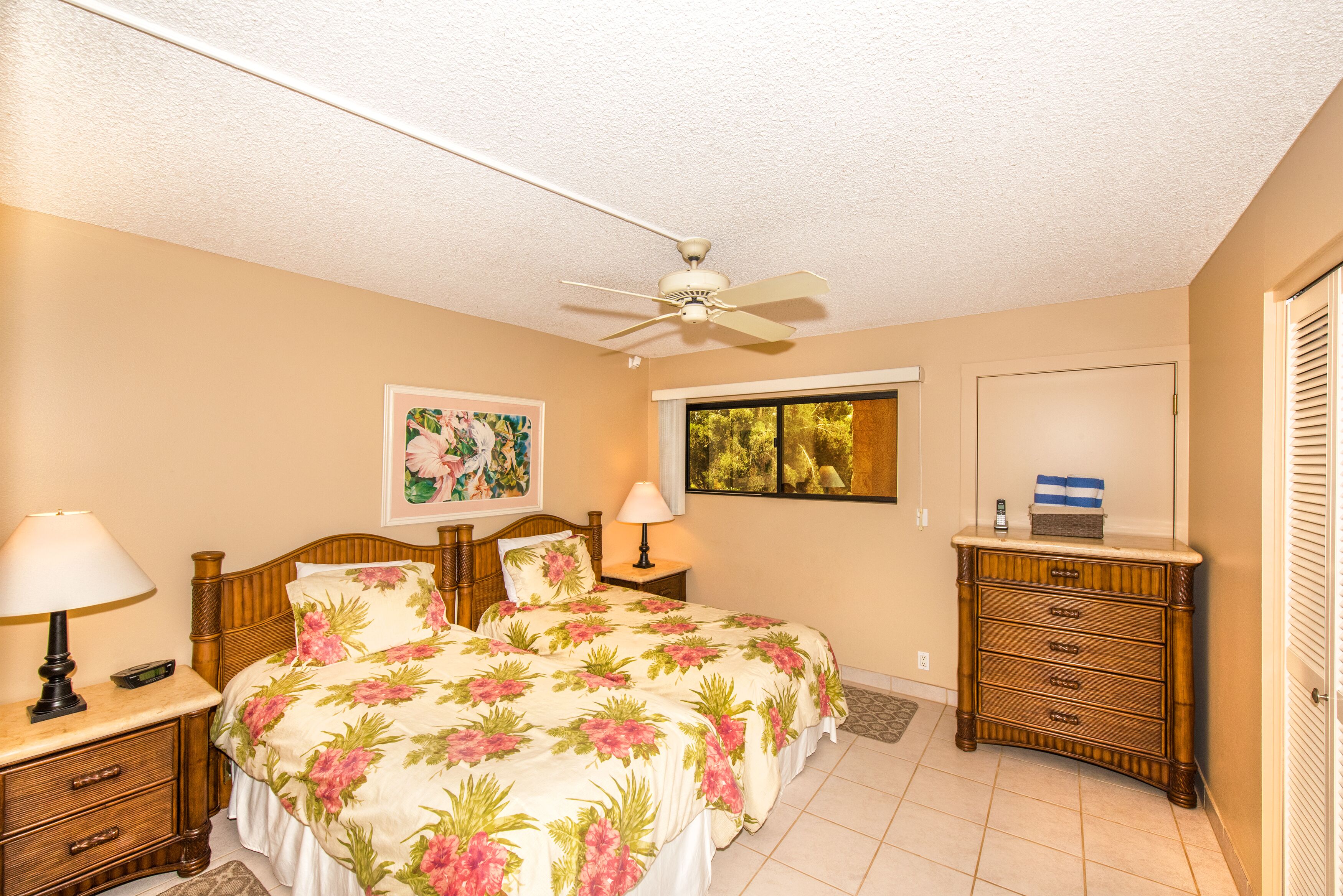 A bedroom with two beds, a ceiling fan, a nightstand with a lamp, a dresser, and artwork on the walls.