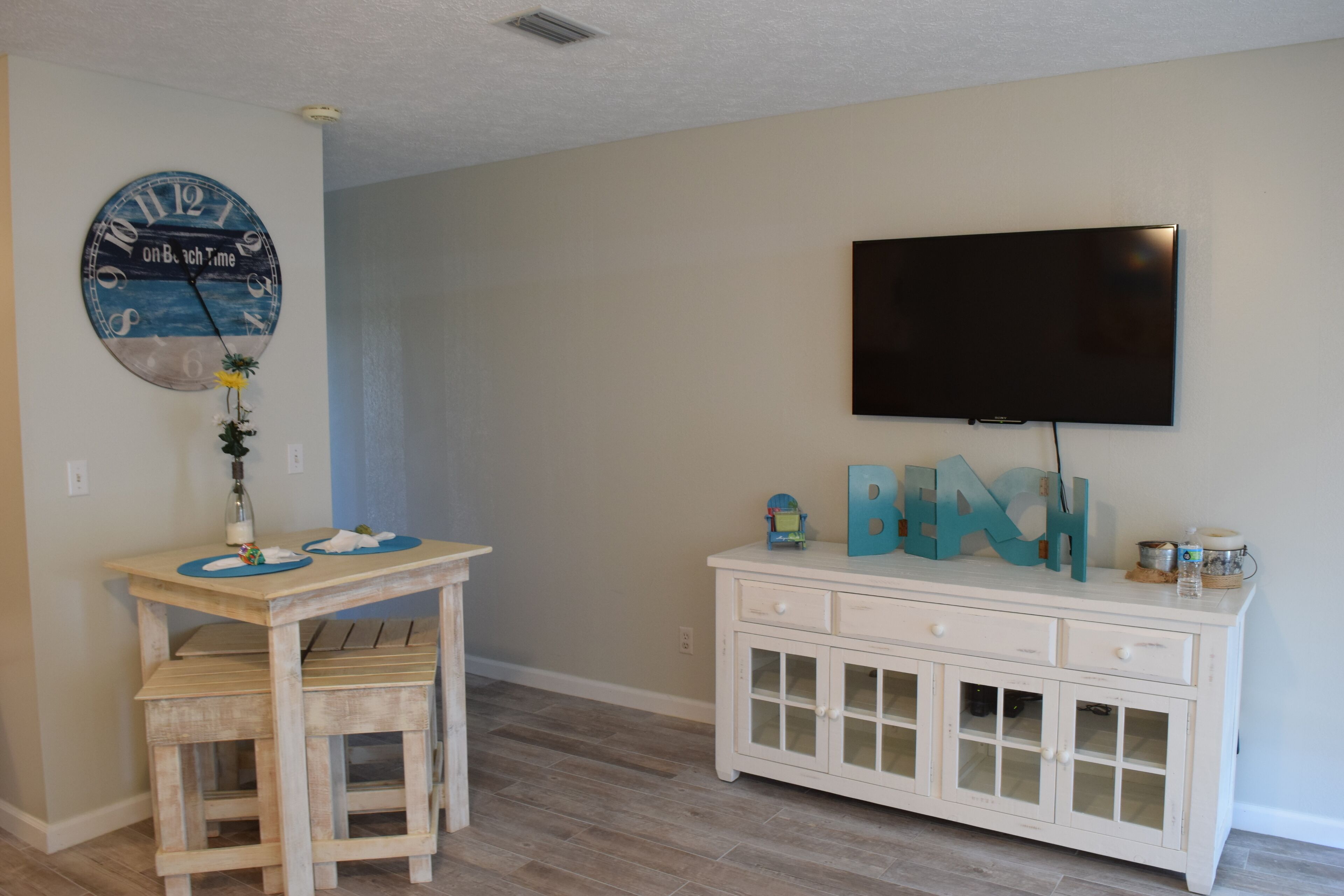 A room with a wooden table, a white cabinet, a clock, and a television mounted on the wall.