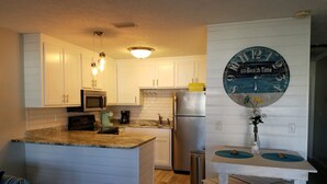 A kitchen with white cabinets, a stainless steel refrigerator, a microwave, and a coffee maker. There is a small island with a sink and a clock on the wall.