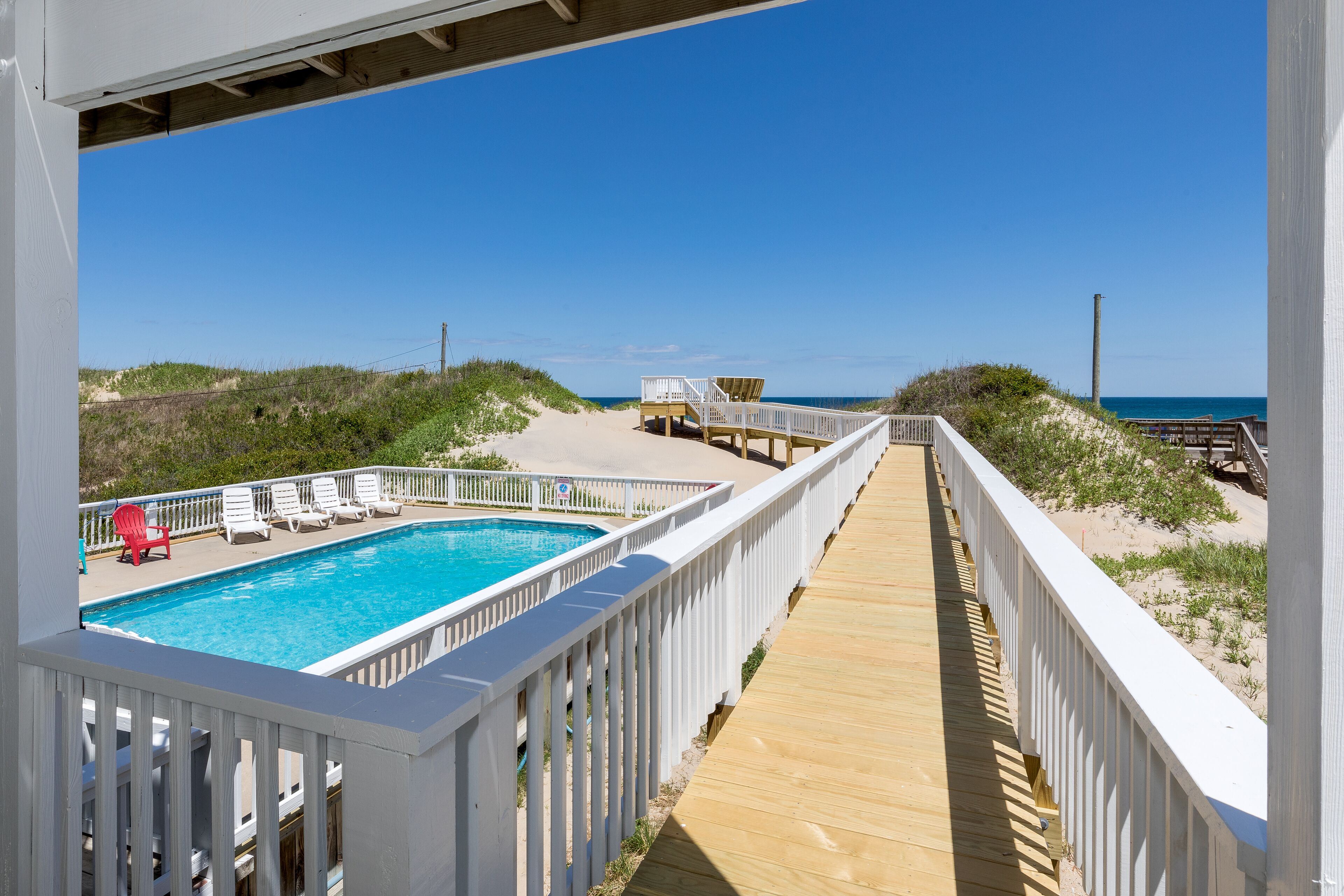 Une terrasse en bois menant à une piscine, avec des rampes blanches, entourée de végétation côtière et surplombée par un ciel bleu clair.