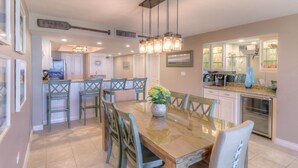 A dining area with a wooden table, chairs, and a vase of flowers. There is a kitchen area with cabinets and a refrigerator in the background.