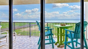 A balcony with a view of the ocean, green chairs, a small table, and a bench.
