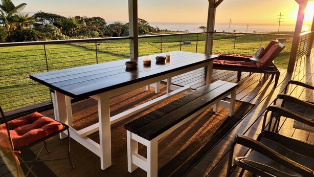Une terrasse extérieure avec une table et des bancs, donnant sur une pelouse et offrant une vue sur l’océan au coucher du soleil.