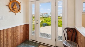 A hallway with a wooden wall panel, a door with a view to the outside, and a chair with a woven seat. - 5690- Brown Pelican - Spacious Oceanfront Nags Head Retreat (Nags Head)