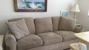 A beige sofa with three cushions, a small white table with a starfish, and a painting of a beach scene on the wall.