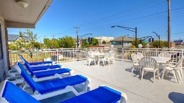 A covered outdoor area with lounge chairs and tables, a white fence, and buildings in the background.