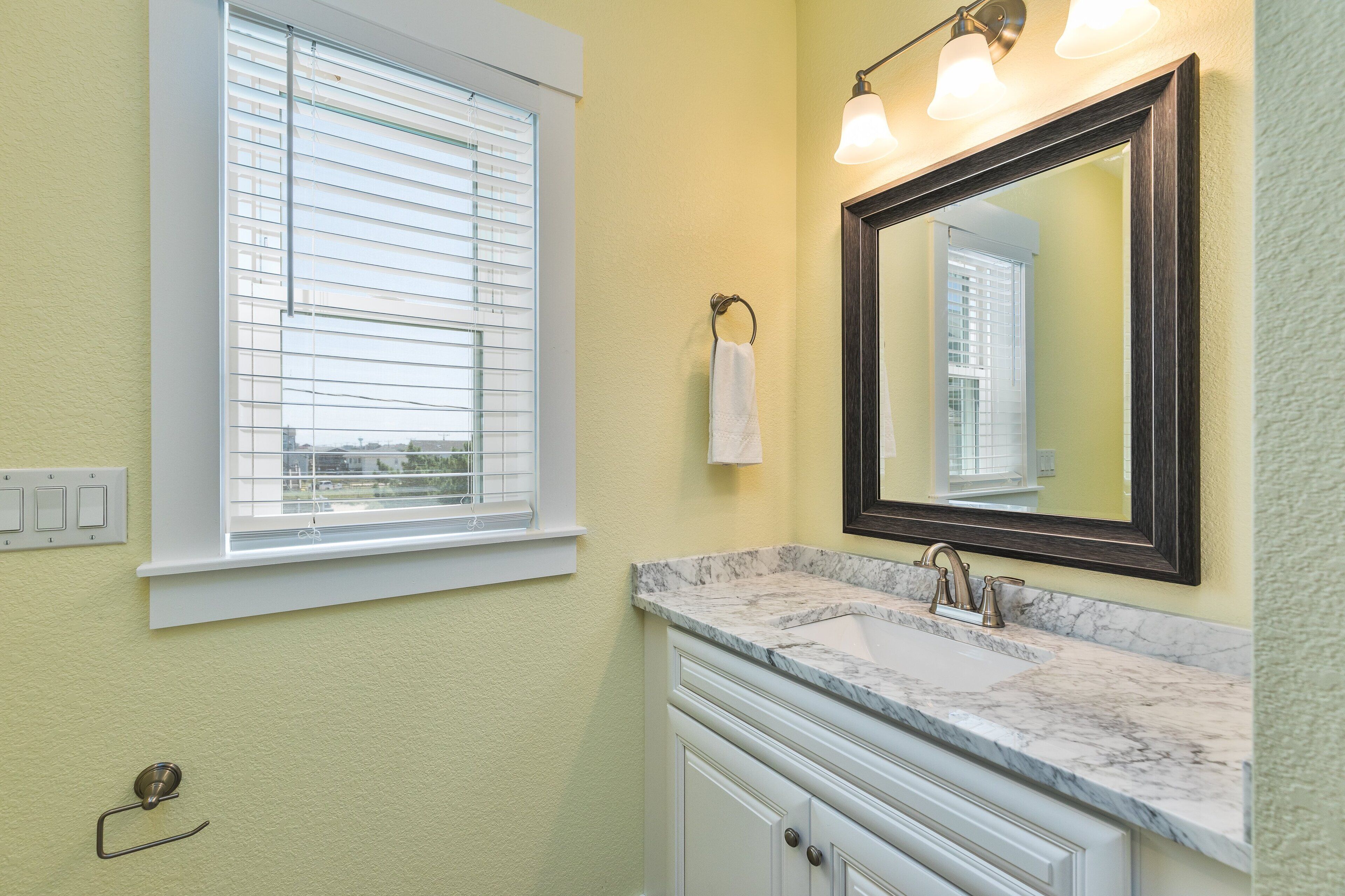 A bathroom with a window, a sink with a marble countertop, and a mirror with lights.