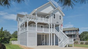 Ein zweistöckiges Strandhaus mit Balkon, einer überdachten Veranda und einer Treppe, die zum oberen Stockwerk führt.