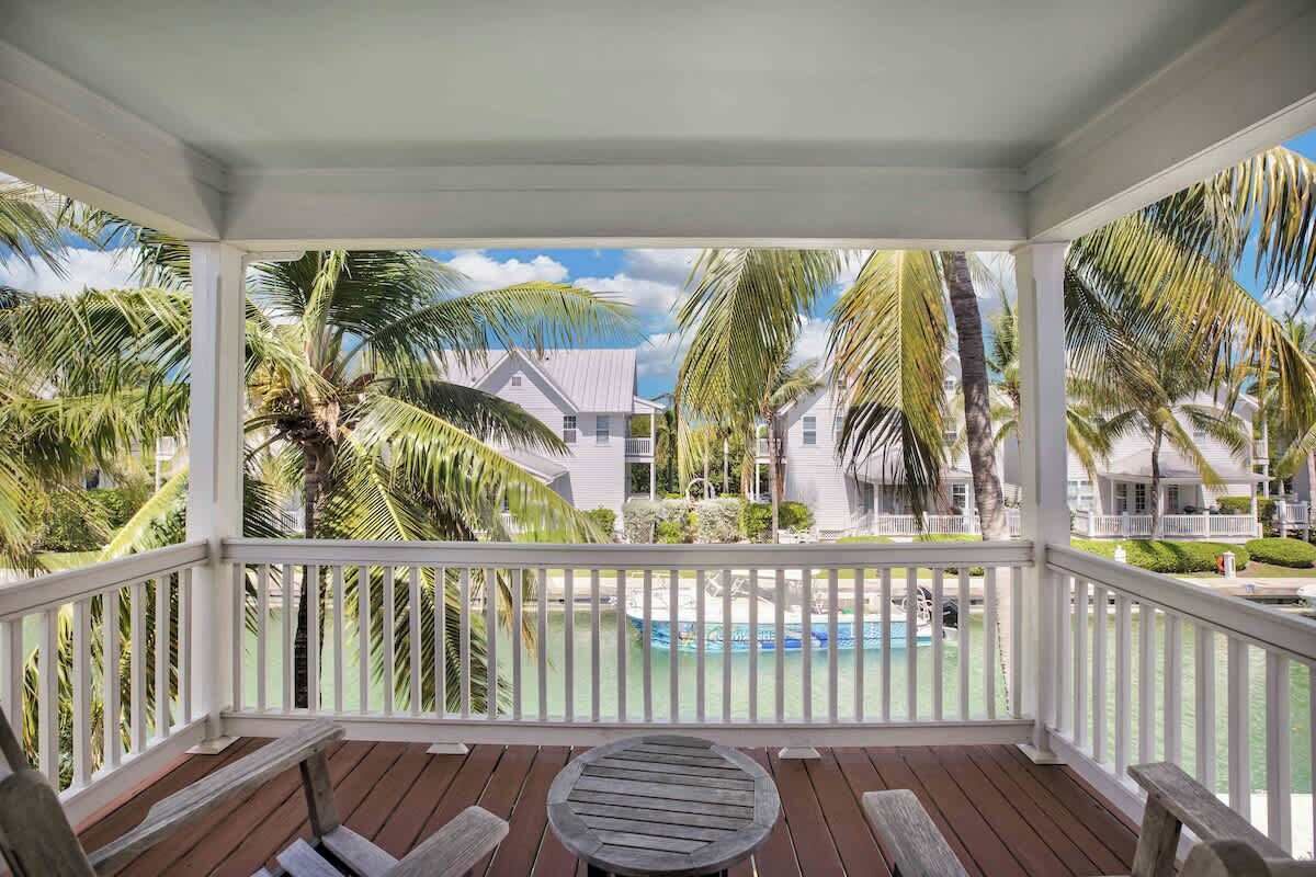A covered outdoor area with a wooden deck, white railings, and a view of a swimming pool surrounded by palm trees and white buildings.