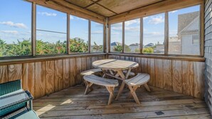 Een overdekt buitengebied met een houten tafel en banken, grote ramen en uitzicht op groen en gebouwen.