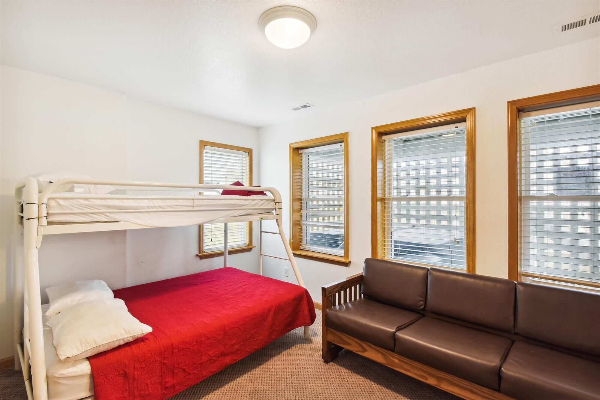 A room with a bunk bed, a red bedspread, a brown leather sofa, and three windows with blinds.