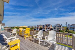 A wooden deck with white and yellow Adirondack chairs, overlooking a row of houses and the ocean.