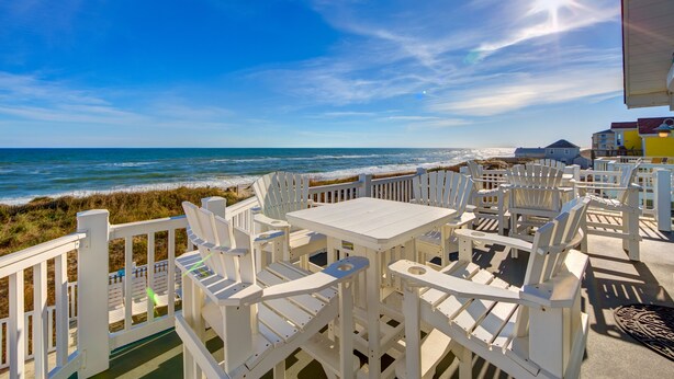 A beachside patio with white Adirondack chairs and a table, overlooking the ocean and beach.