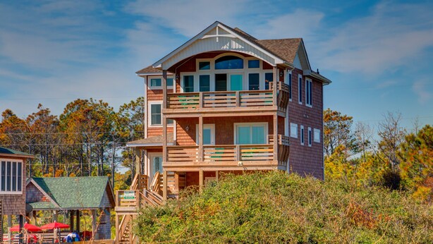 A multi-story wooden building with balconies, a covered porch, and a staircase leading to the upper levels. - 5728- Wench's Pad - Semi-Oceanfront Nags Head | Pool & Hot Tub (Nags Head)