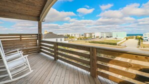 A wooden deck with a white rocking chair, overlooking a beach and buildings. - 5728- Wench's Pad - Semi-Oceanfront Nags Head | Pool & Hot Tub (Nags Head)