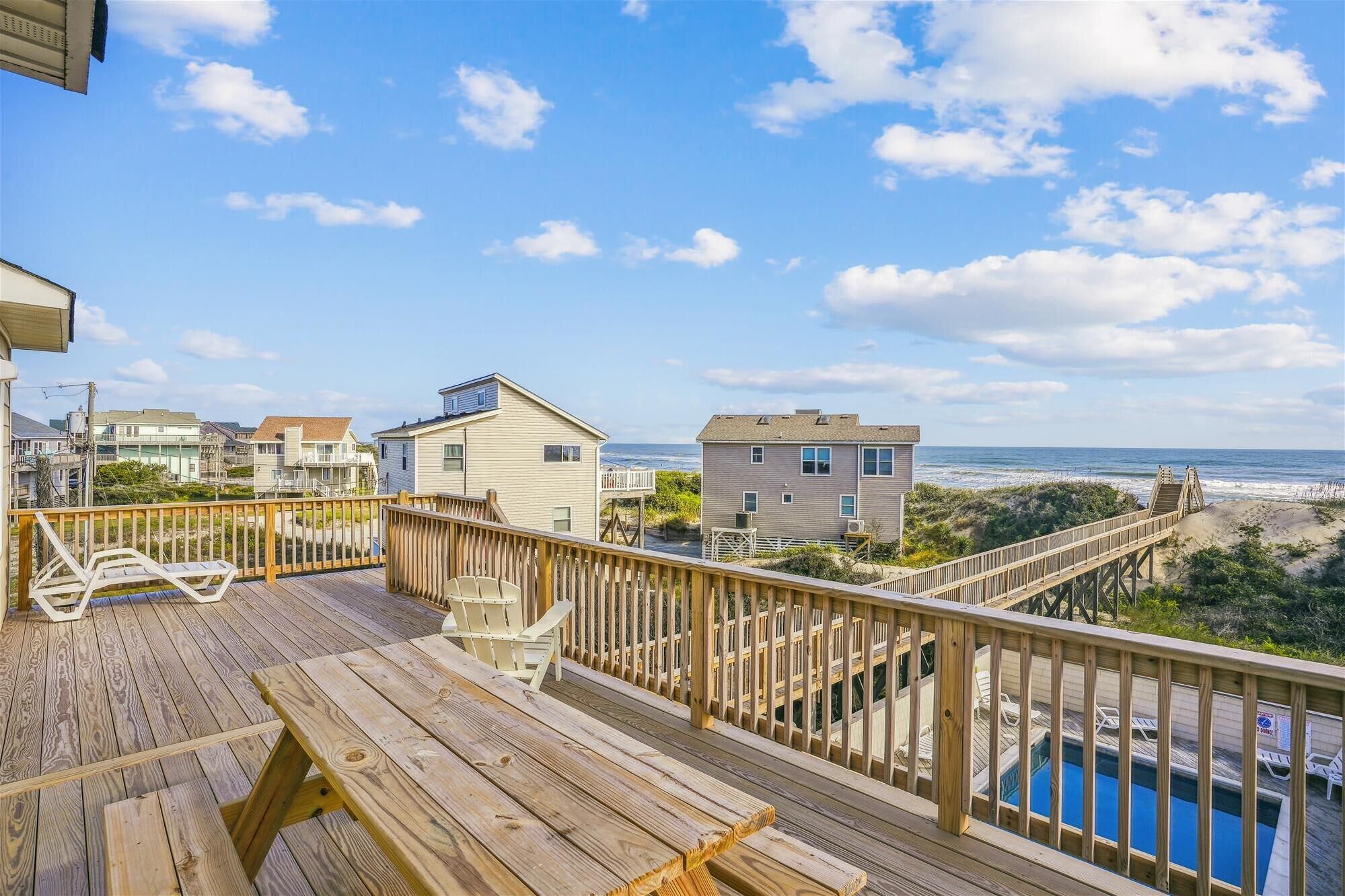 A wooden deck with a pool, overlooking a beach with houses and the ocean.