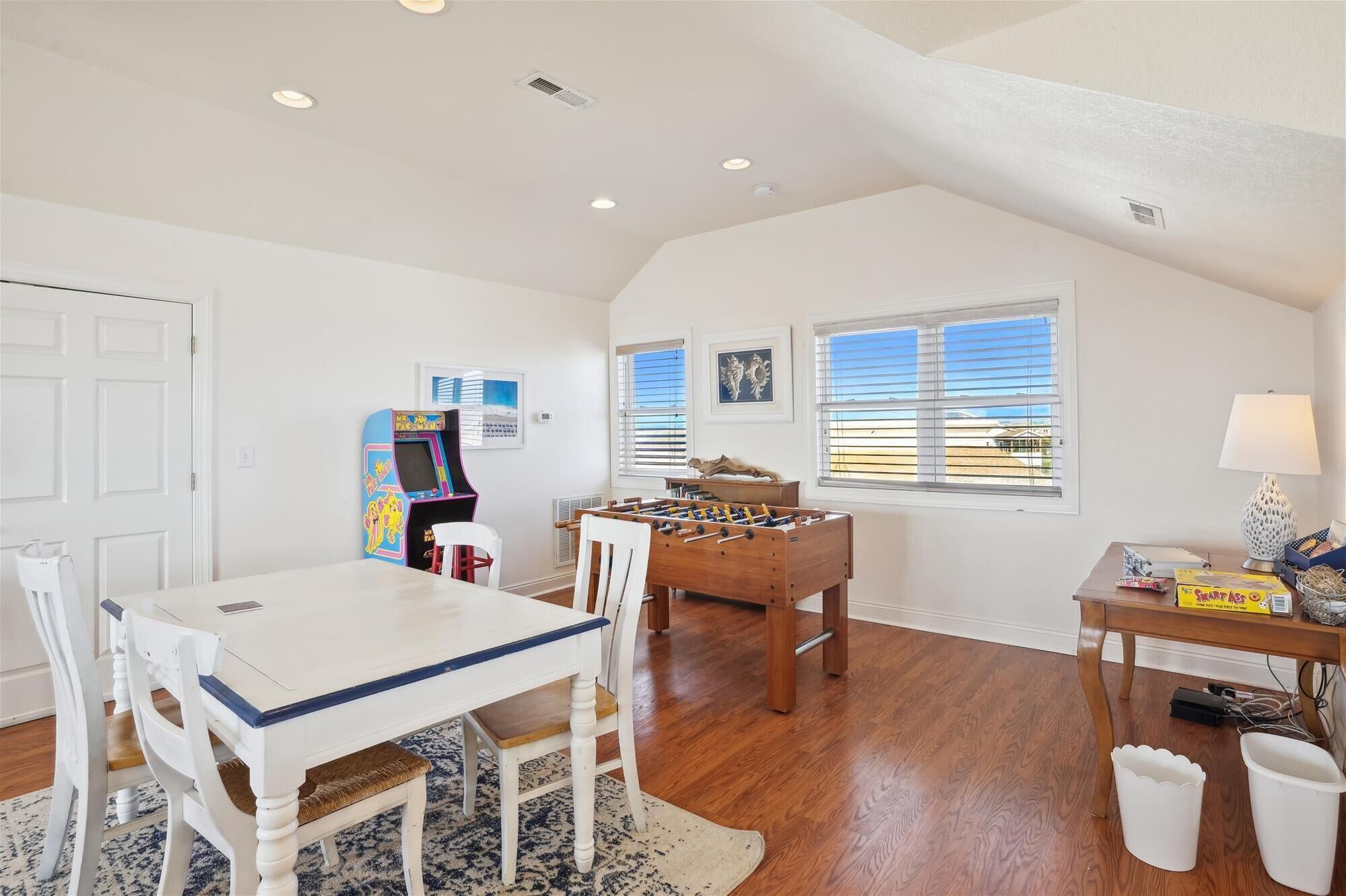 A spacious room with a wooden floor, a white table with chairs, a foosball table, a foosball table, and a window with blinds.