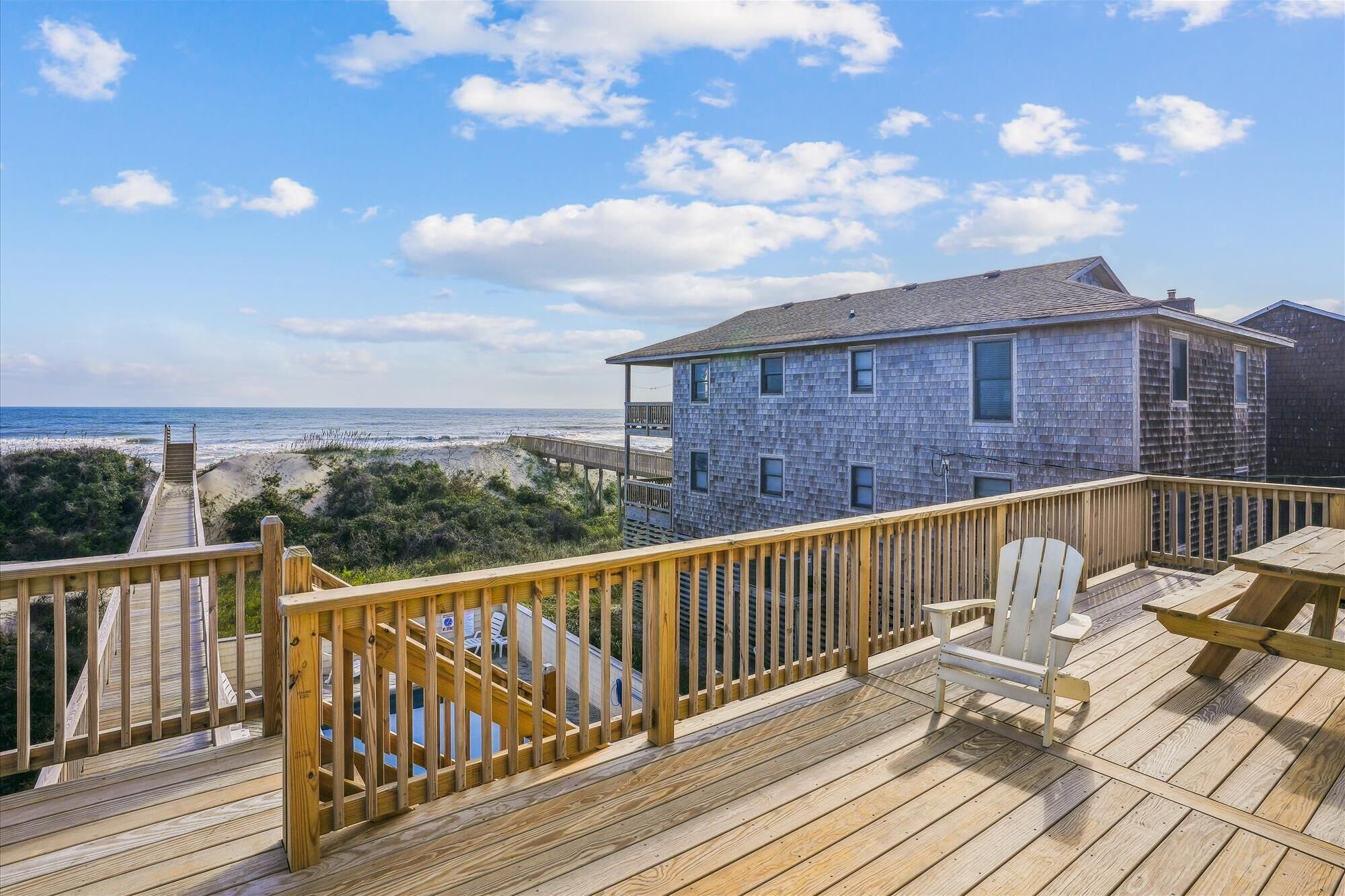 A wooden deck with a view of the beach and ocean, a house with a balcony, and a chair.