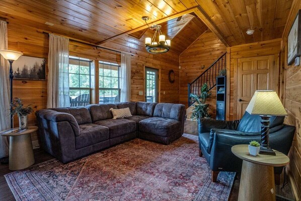 A cozy cabin interior with a large sectional sofa, a small round table, a staircase, and a chandelier. - Chateau: 3 bedroom cabin with hot tub, pool table, lake view (Himrod)