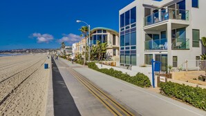 Ein Küstenhotel im modernen Design, mit Balkonen und Glasgeländern, einem gepflasterten Weg und Blick auf den Strand.