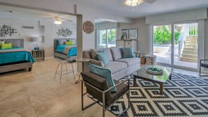 A modern living room with a sofa, coffee table, and a dining area with a chair and table. There is a ceiling fan, a large window with a view of the outdoors, and a patterned rug on the floor.
