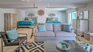 A modern living room with a sofa, armchair, and coffee table. There is a bed with blue bedding in the background.
