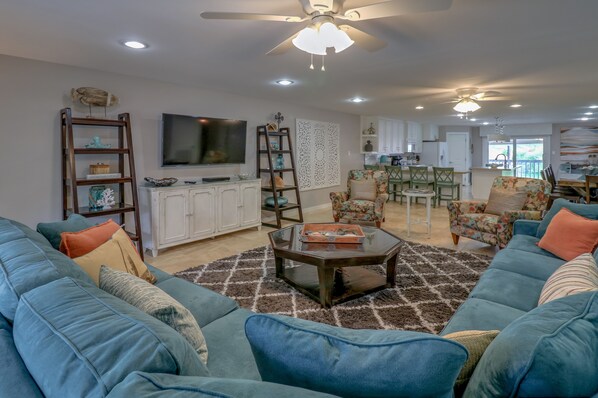 A spacious living room with a large sofa set, a coffee table, and a TV. There is a kitchen in the background with a dining area.