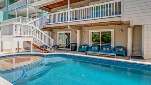 A swimming pool with a white deck, blue chairs, and a wooden staircase leading to a balcony.