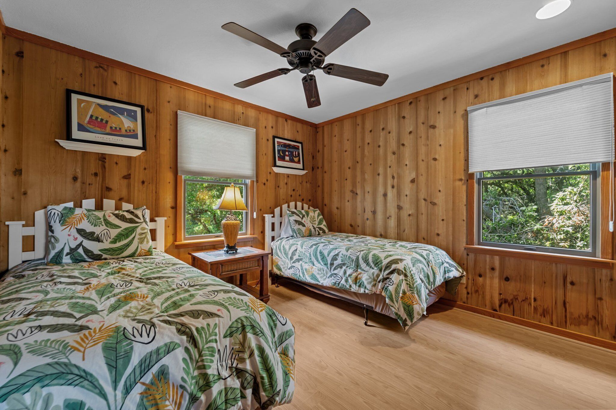 A room with two beds, wooden walls, a ceiling fan, and a window with a view of greenery.