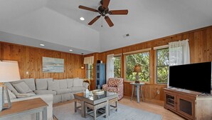 Una sala de estar con pared de madera, ventilador de techo, ventana grande con cortinas, sofá, sillón estampado, mesa de centro, televisor sobre un mueble de madera y una lámpara.