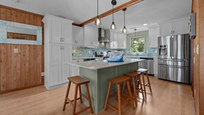 Cocina moderna con armarios blancos, una isla central con taburetes, electrodomésticos de acero inoxidable e iluminación colgante.