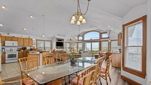 A spacious kitchen with wooden cabinets, a large island, and a dining area with a glass table and wicker chairs. - 5785- Southern Comfort - South Nags Head Oceanfront Retreat (Nags Head)