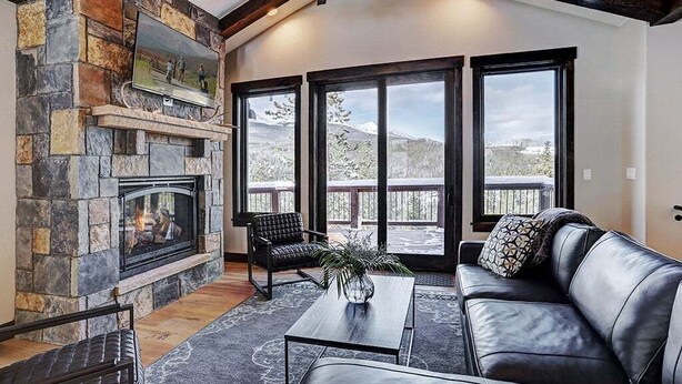 A cozy living room with a stone fireplace, leather sofa, and large windows offering a view of the outdoors.