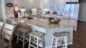 A kitchen with white cabinets, a granite countertop, a sink, a microwave, a refrigerator, and a sign that reads "Beach Bungalow".