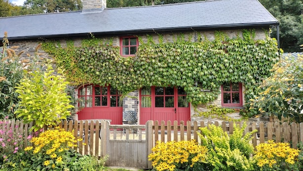 A stone house with red doors and windows, surrounded by a wooden fence and various flowering plants. - A characterful former Carriage House, located in peaceful scenic countryside (Cellan)